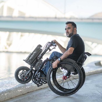 Handbike BATEC MINI 2 de color gris grafito plegado y anclado a una silla de ruedas manual, destacando su motor eléctrico.