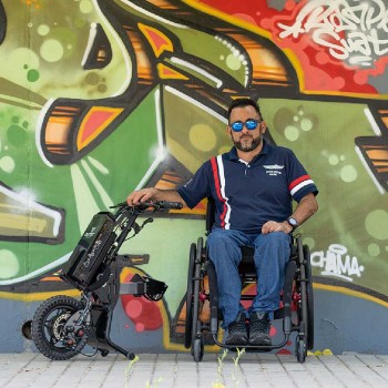 Handbike BATEC MINI 2 de color gris grafito plegado y anclado a una silla de ruedas manual, destacando su motor eléctrico.
