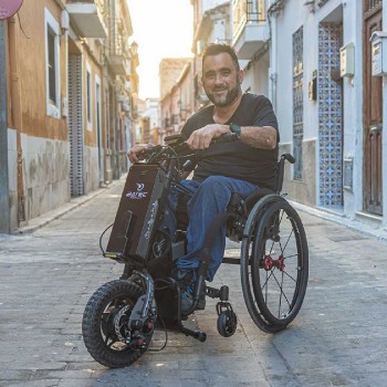 Handbike BATEC MINI 2 de color gris grafito plegado y anclado a una silla de ruedas manual, destacando su motor eléctrico.